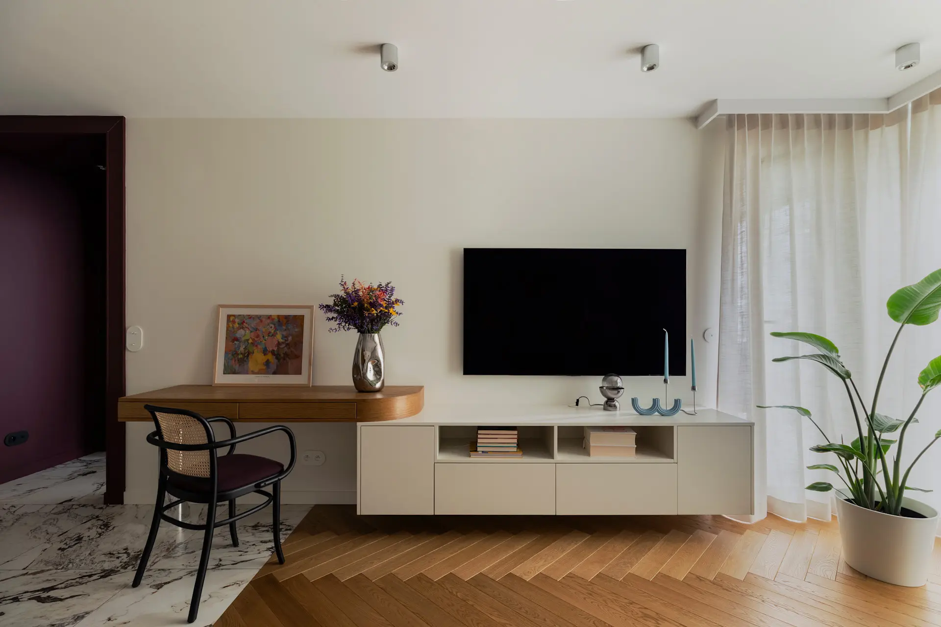Nowoczesna aranżacja salonu z białą szafką RTV, drewnianym biurkiem, krzesłem rattanowym i rośliną doniczkową przy oknie z lnianymi zasłonami.