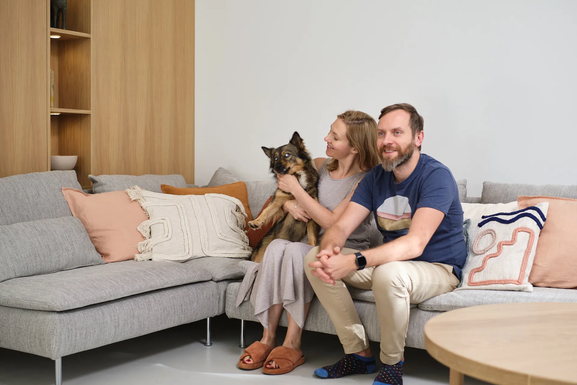 Nowoczesny salon z szarą narożną sofą, drewnianą zabudową ścienną z półkami, dodatkami w brzoskwiniowych odcieniach i okrągłym stolikiem kawowym.