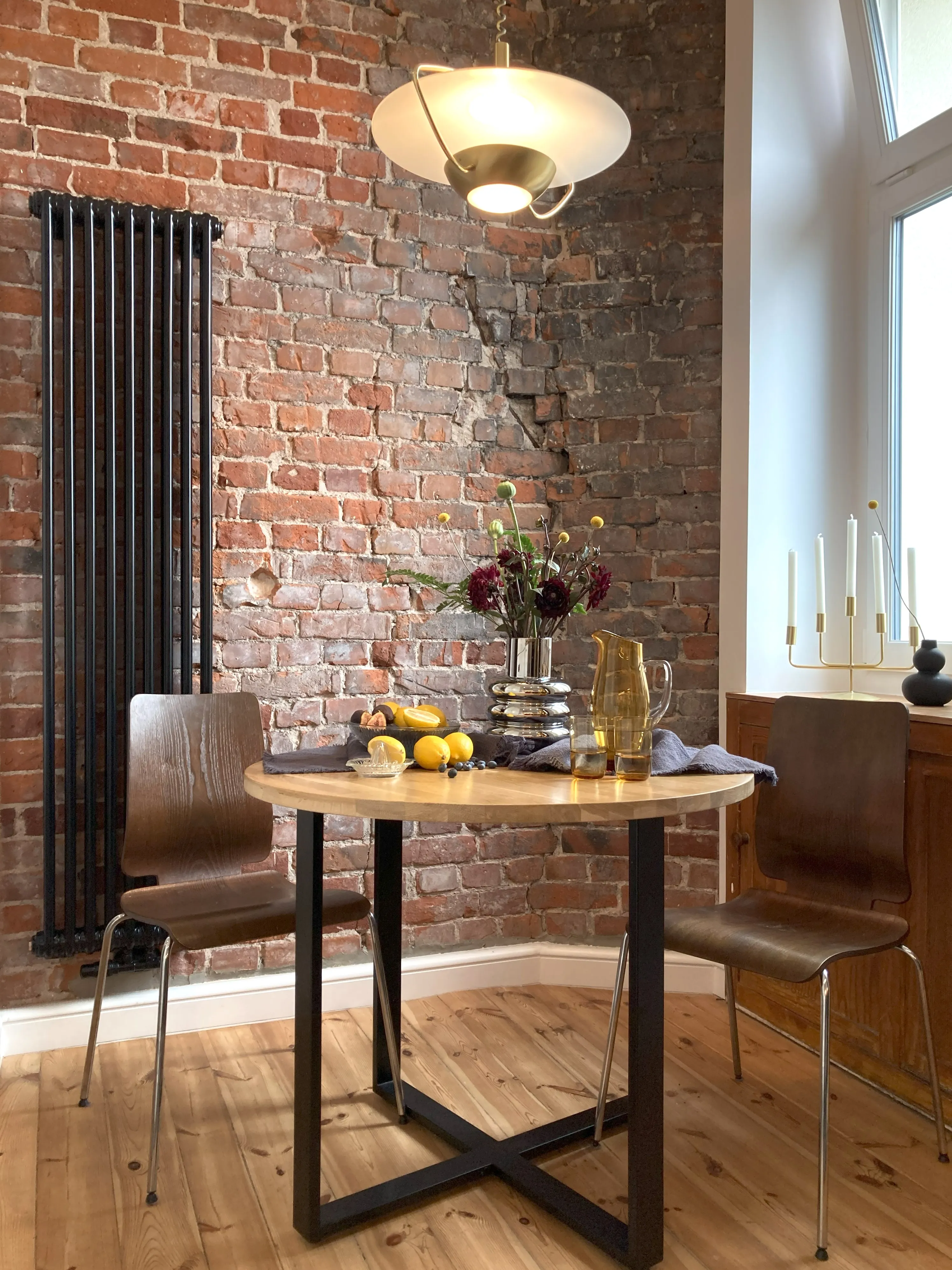 Aranżacja jadalni w stylu loft z okrągłym drewnianym stołem, brązowymi krzesłami, ceglaną ścianą, czarnym grzejnikiem i nowoczesną złotą lampą wisząca.