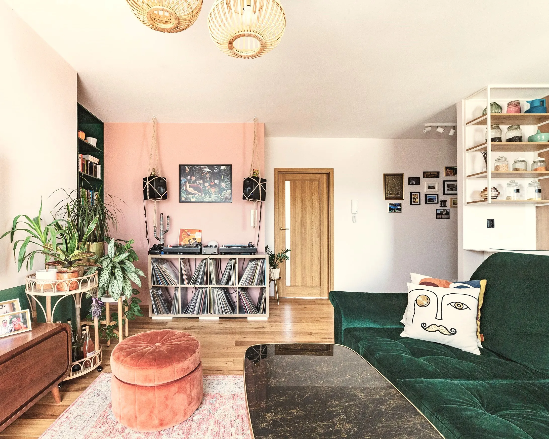 Nowoczesny salon z zielonym aksamitnym sofą, różową ścianą akcentową, drewnianym regałem na płyty, pufą w odcieniu koralowym i naturalnymi roślinami — przykład aranżacji wnętrz łączący retro z współczesnym designem.