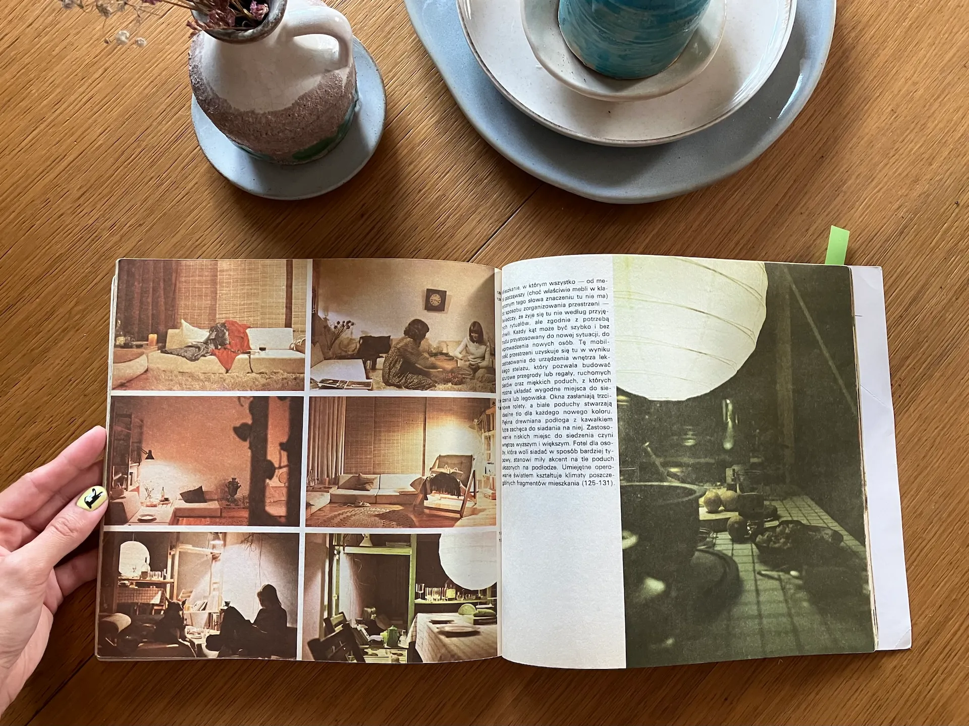 Otwarta książka o projektowaniu wnętrz z archiwalnymi fotografiami pomieszczeń na drewnianym stole, obok ceramicznych naczyń w bieli i turkusie.