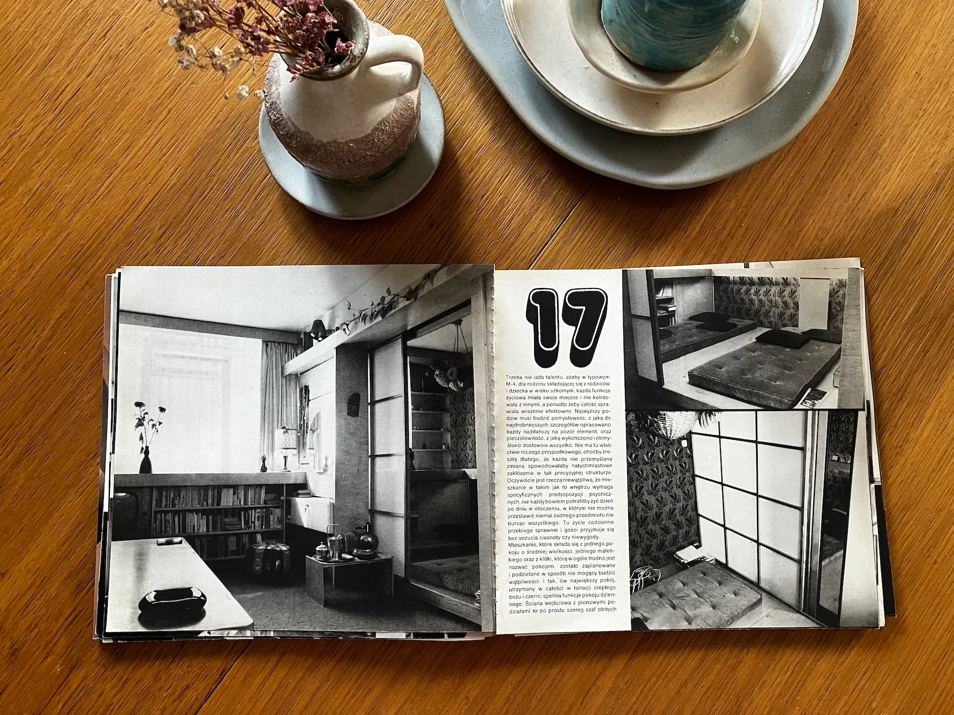 Otwarty magazyn o projektowaniu wnętrz leży na drewnianym blacie obok białych naczyń z suszonymi kwiatami; strony prezentują czarno-białe fotografie projektu sypialni i salonu w stylu minimalistycznym.