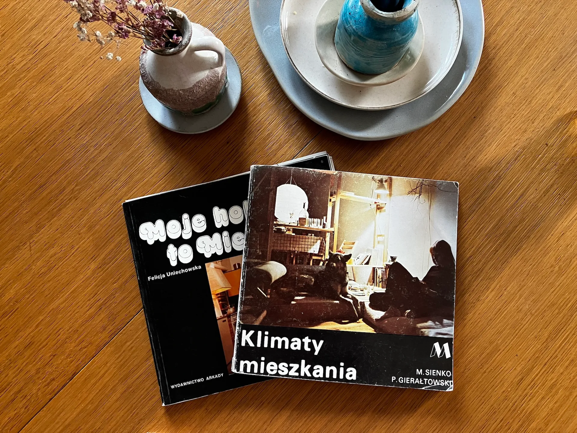 Drewniana powierzchnia stołu z projektami wnętrz, ceramicznymi wazonami w odcieniach błękitu i beżu oraz albumami o aranżacji pomieszczeń, prezentujące inspiracje do projektowania nowoczesnych wnętrz.