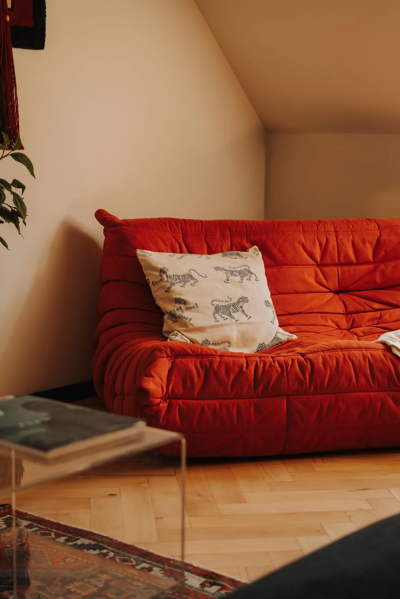 Czerwona pikowana sofa w minimalistycznym wnętrzu na poddaszu z beżowymi ścianami, drewnianą podłogą i dekoracyjną poduszką w zwierzęce wzory.
