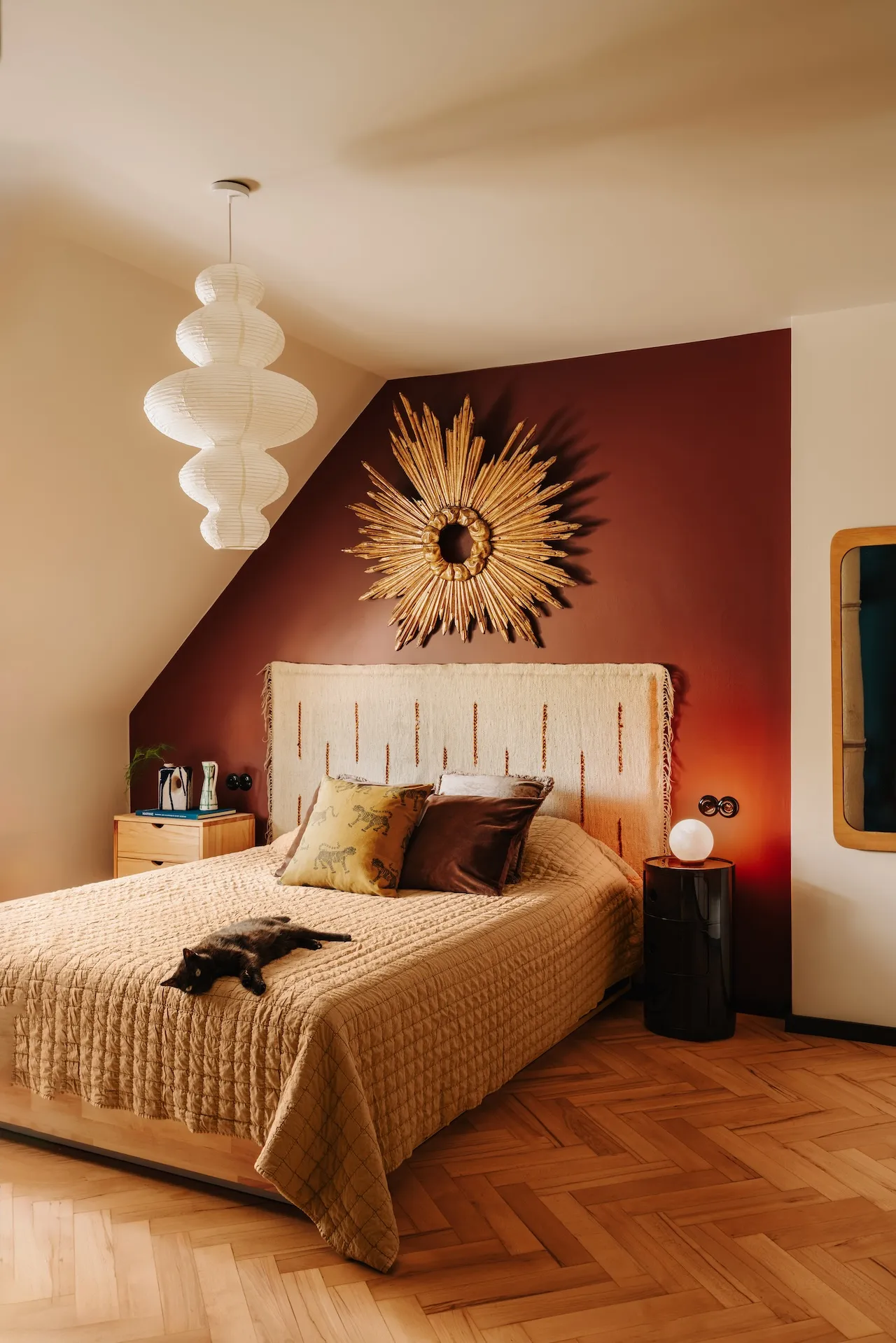 Sypialnia na poddaszu z burgundową ścianą, łóżkiem z beżowym zagłówkiem, białą designerską lampą i złotym dekoracyjnym słońcem, aranżacja w ciepłych tonach ochry i terrakoty.