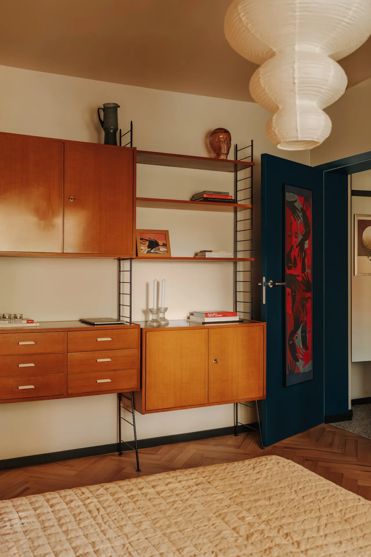 Vintage aranżacja sypialni z drewnianymi meblami lat 60., modularnymi regałami, brązowym sufitem i ciemnoniebieskimi drzwiami, na tle beżowych ścian i drewnianej podłogi.
