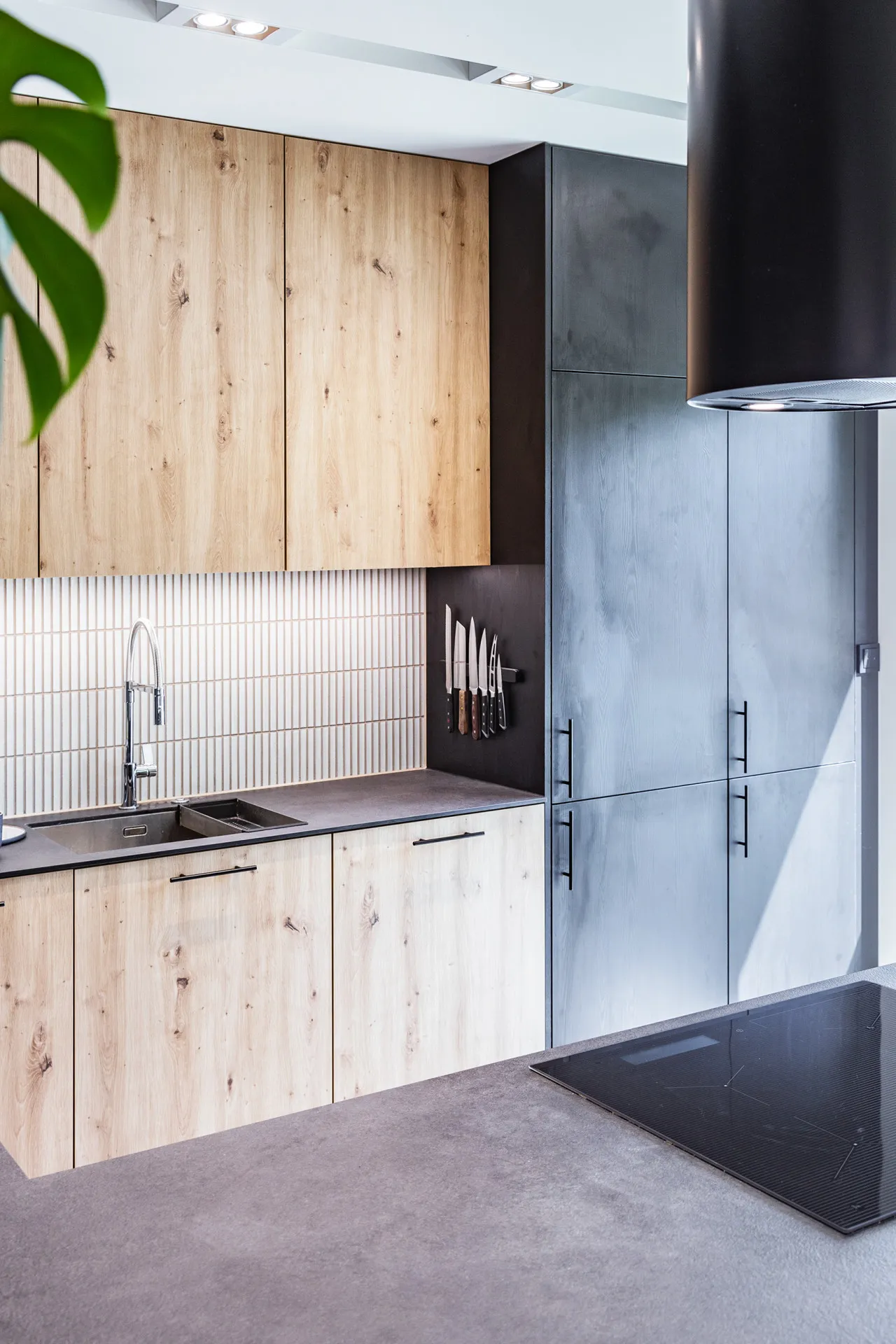 Nowoczesna kuchnia z drewnianymi frontami, białą płytką w ryflowanie, czarnym blatem i stalowymi urządzeniami. Aranżacja łączy naturalne drewno z minimalistycznym designem i industrialnym akcentem – czarną ścianą z wieszakiem na noże.