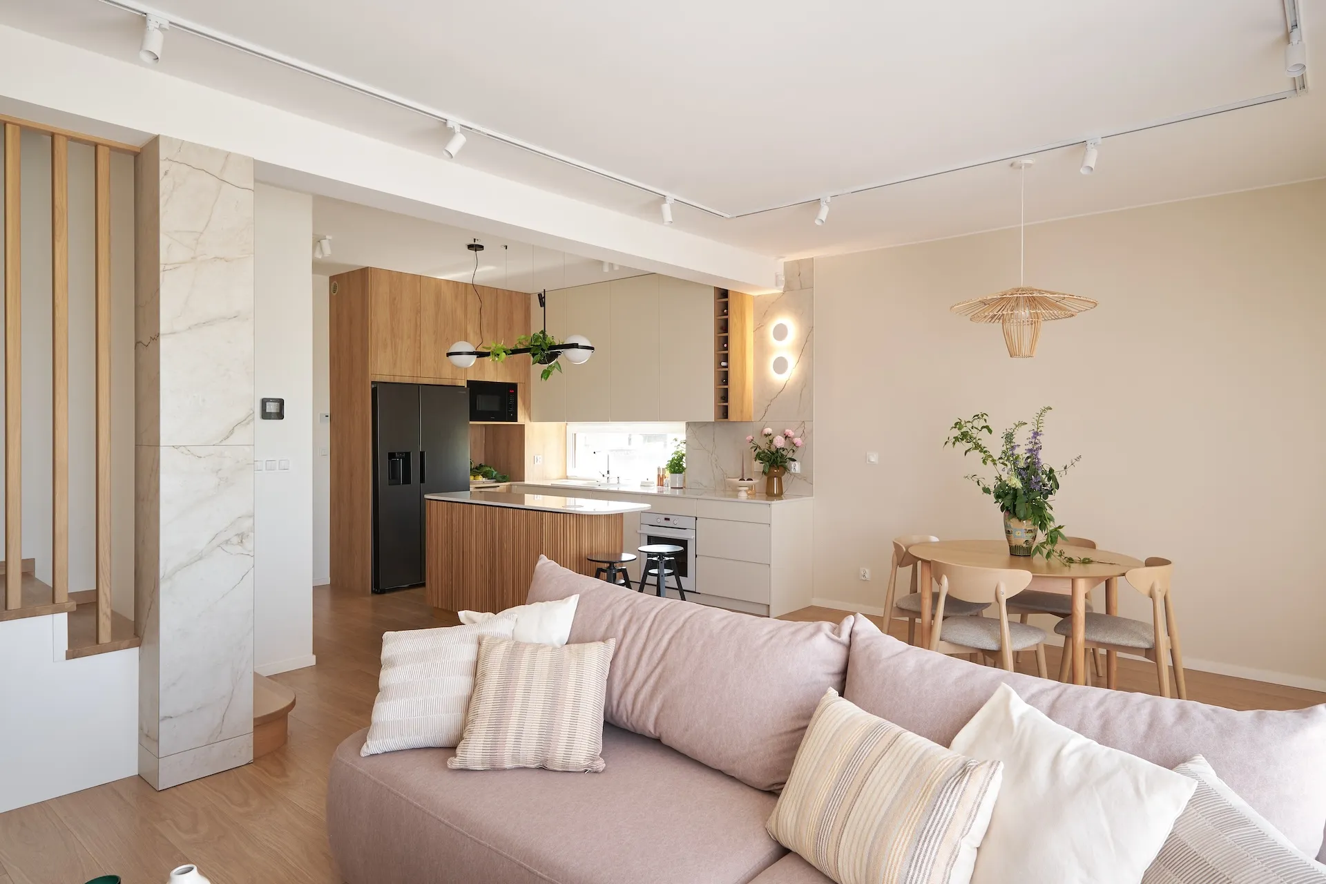 Otwarte wnętrze apartamentu z drewnianą kuchnią, marmurkowym panelem, sofą w odcieniach beżu i różu, drewnianym stołem jadalnym oraz nowoczesnym oświetleniem.