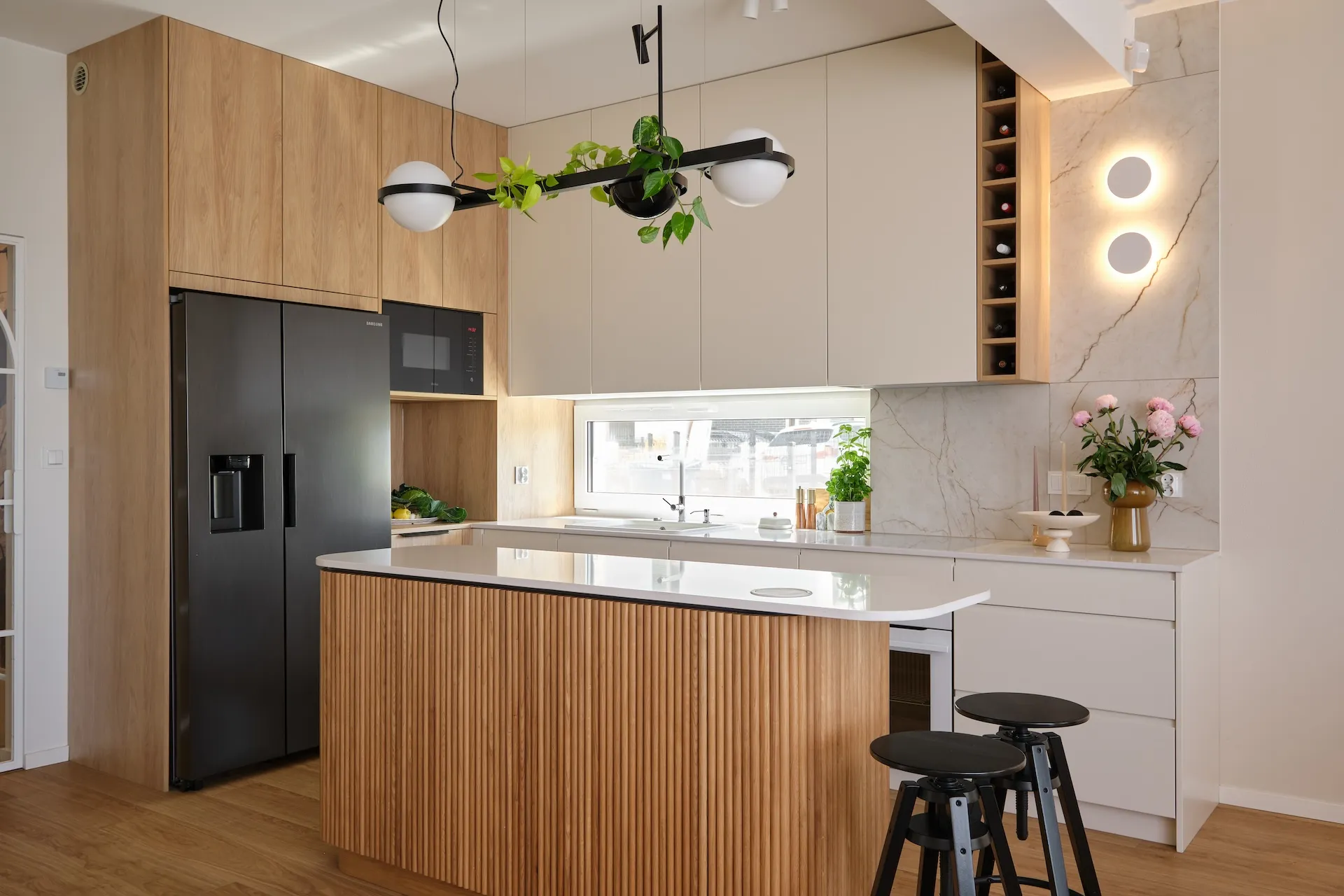 Nowoczesna kuchnia z drewnianym frontem wysepki, marmurowym blatem i białymi szafkami, oświetlona designerską lampa z roślinami i czarnymi hokerami.