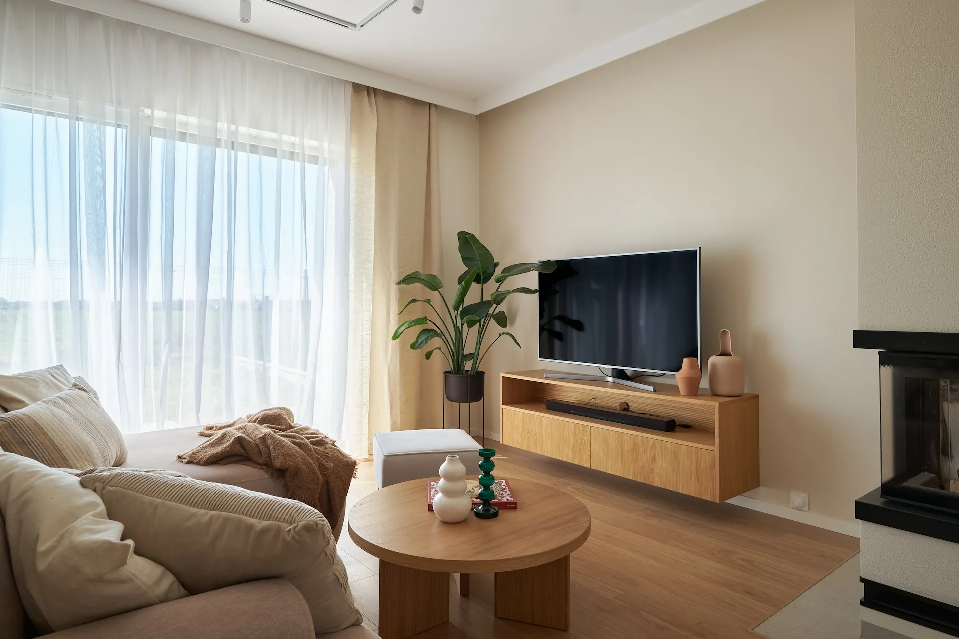 Przytulny salon z beżową kanapą, drewnianym stolikiem, modułową szafką pod telewizor i kominkiem, oświetlony naturalnym światłem przez panoramiczne okna z zasłonami w odcieniach kremowych.
