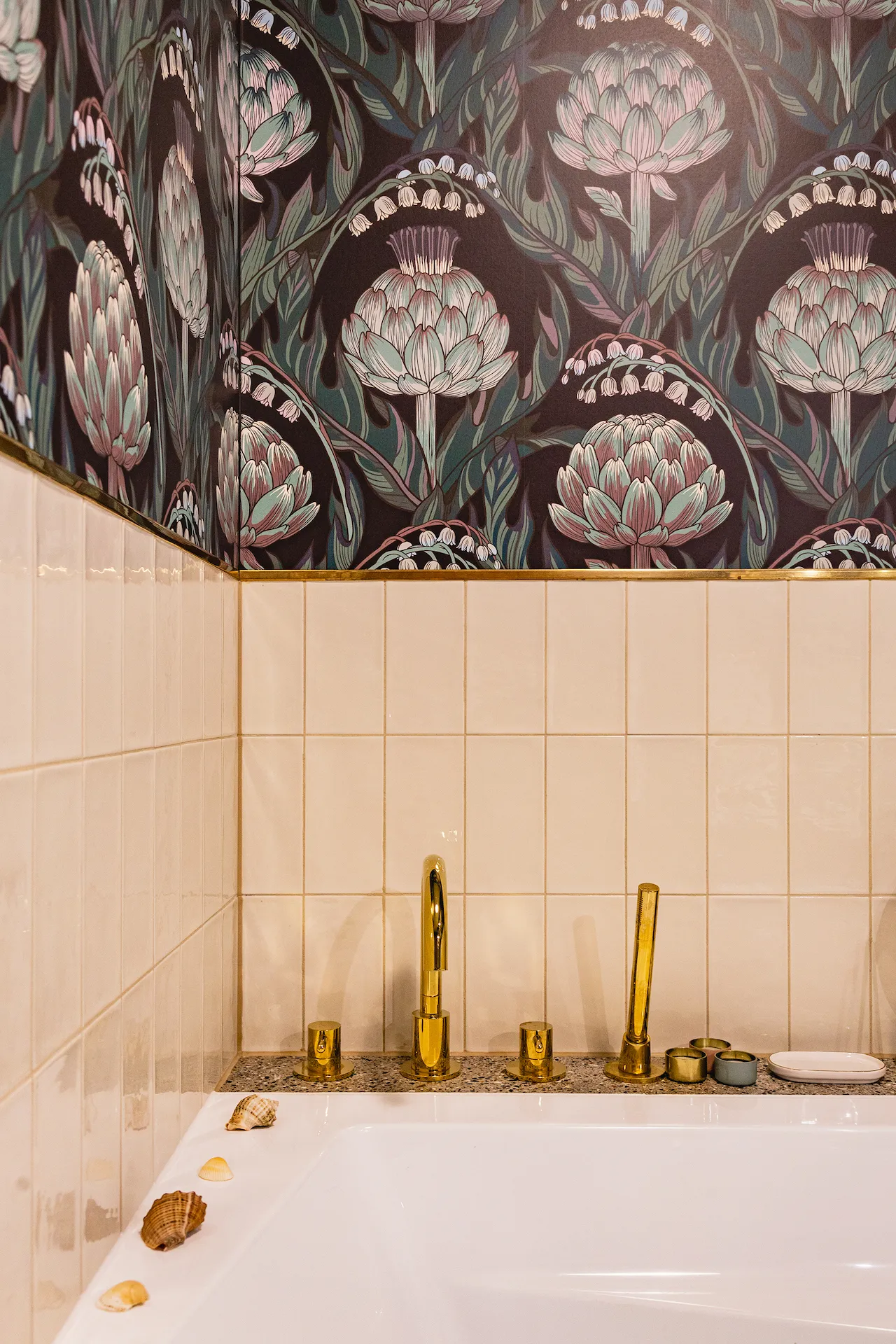 Elegancka łazienka z miodowo-beżową ceramiką, złotymi armaturami i tapetą botaniczną z motywem różowych kwiatów na ciemnym tle, ozdobiona muszlami i naturalnym dekorem.