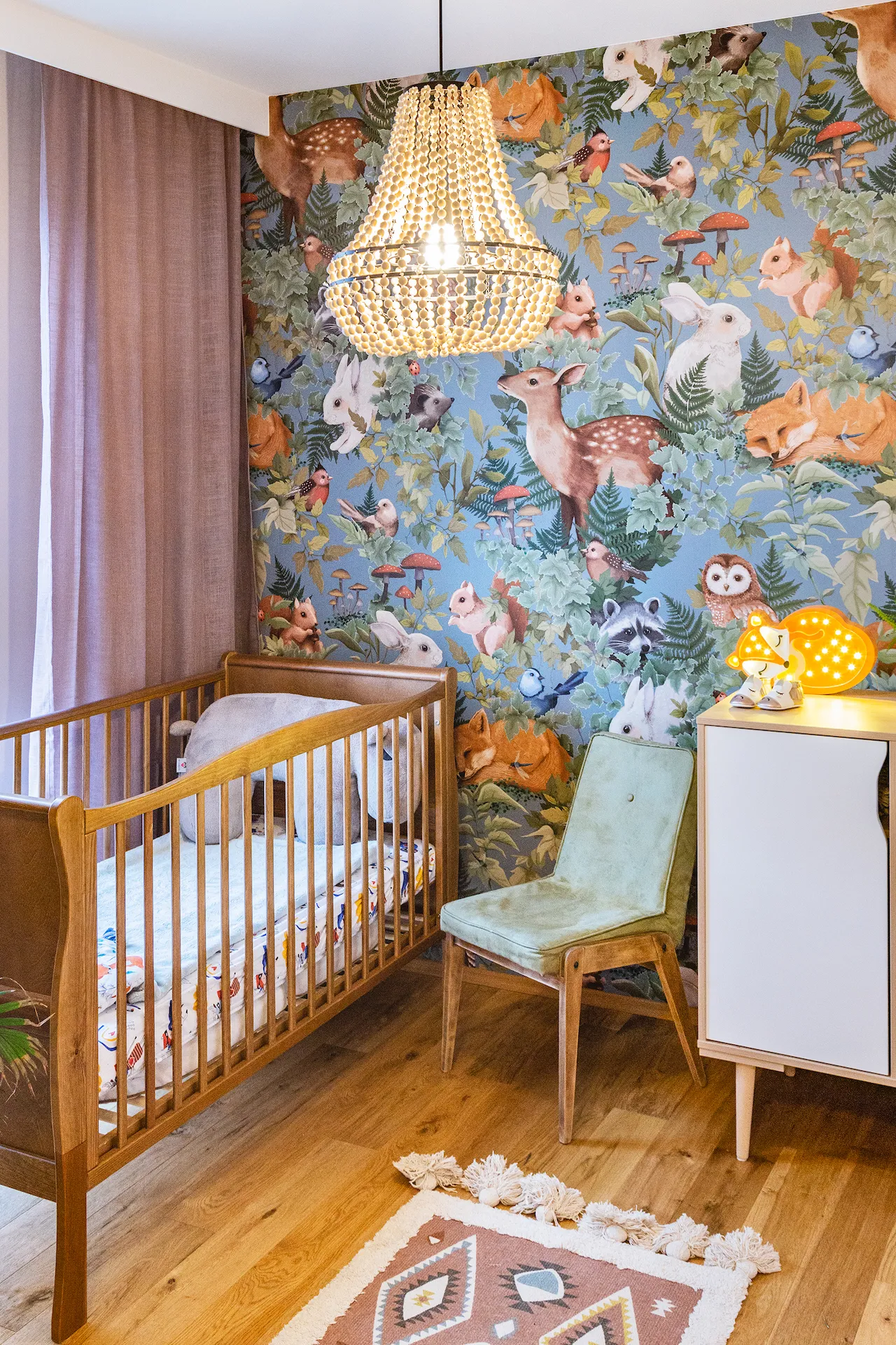 Projektowanie pokoju dziecka z drewnianym łóżeczkiem, tapetą ze zwierzętami w lesie, kryształowym żyrandolem i miętową fotelką retro na drewnianej podłodze.