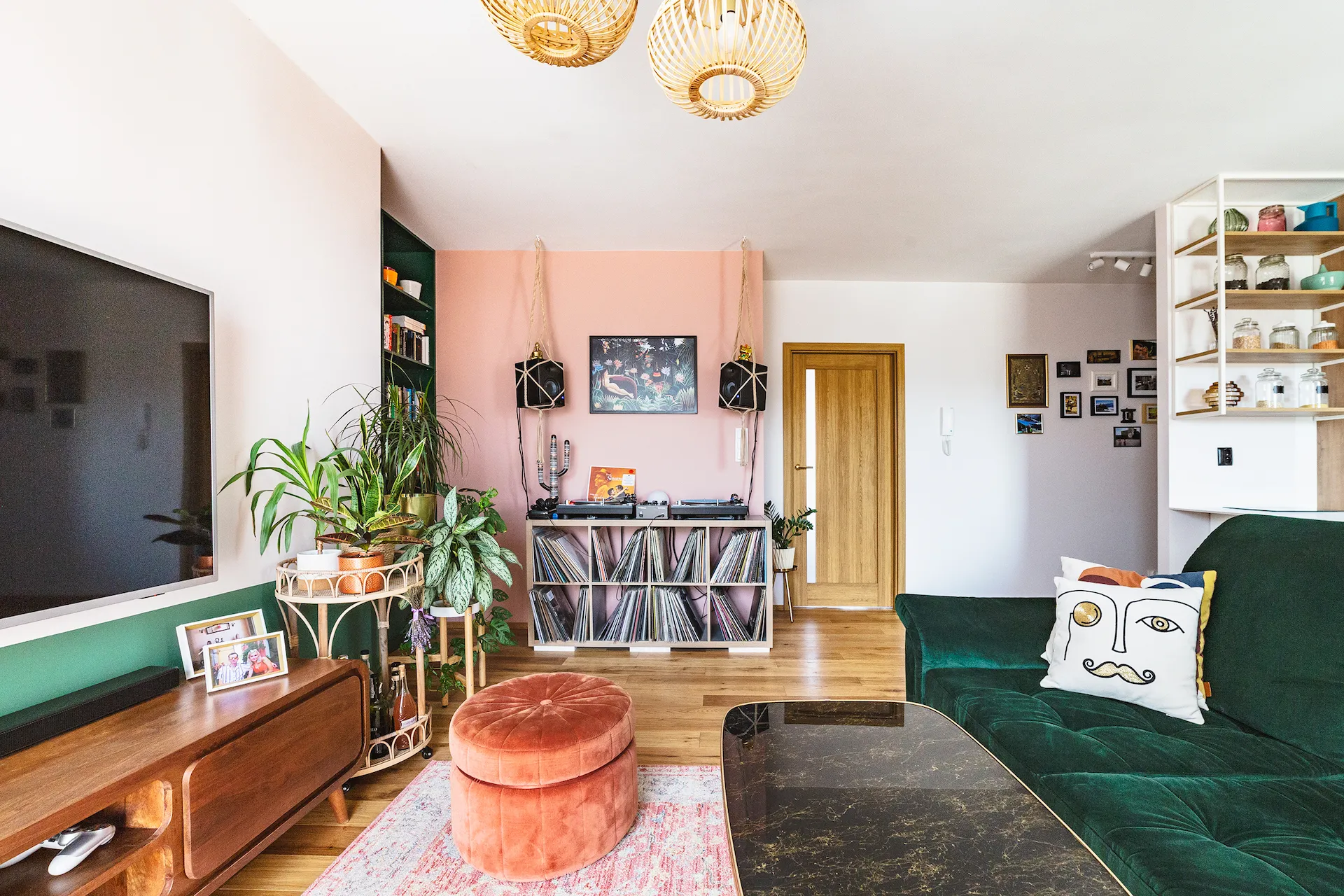 Nowoczesny salon z zielonym welurowym sofą, różową akcentową ścianą, drewnianymi meblami retro i złotymi lamparami sufitowymi w projektowaniu wnętrz Kamiko.