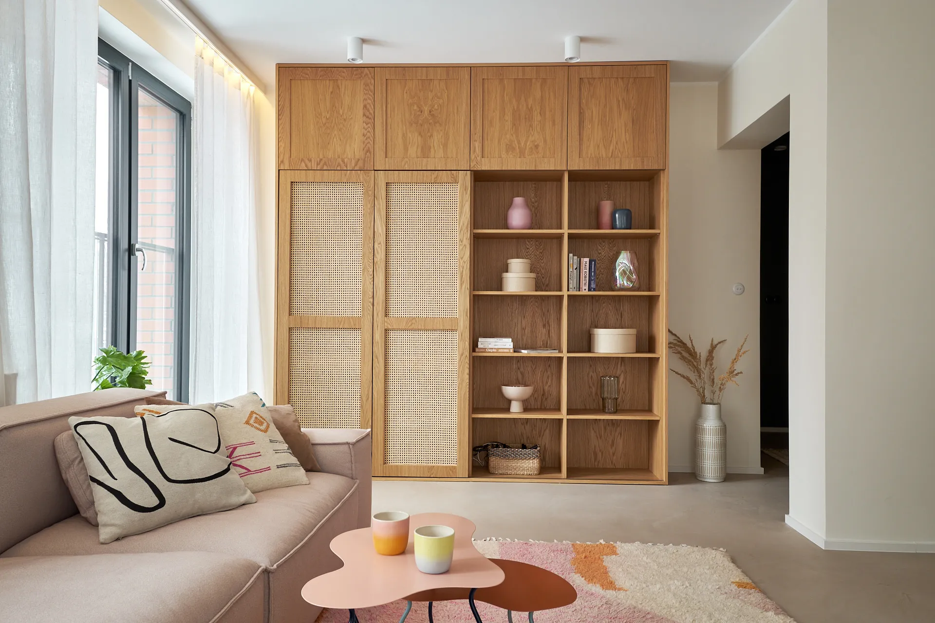 Nowoczesny salon z drewnianą ścianą regałową, beżową sofą i designerskim stolikiem kawowym, oświetlony naturalnym światłem z dużego okna.