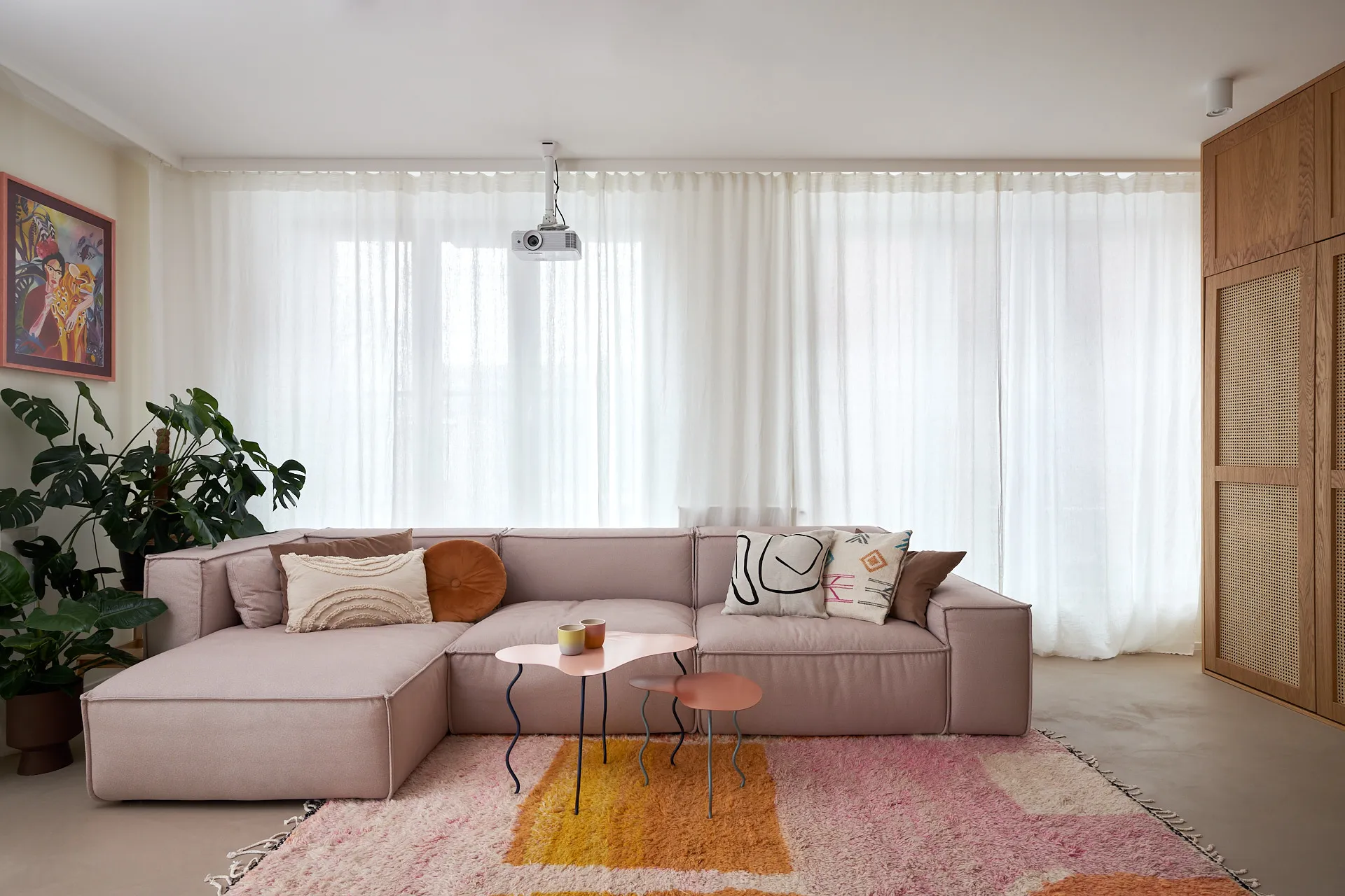 Nowoczesny salon z różową modulową sofą, geometrycznymi poduszkami i stolikami kawowymi na tle dużych okien z białymi zasłonami, drewnianymi drzwiami i kolorowym dywanem.